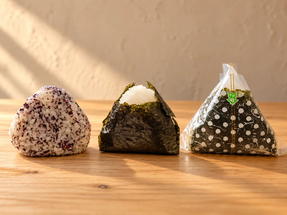 Tre onigiri på ett träbord: en med röd bladmynta i riset, en med sjögräs runt och en i plastfilm